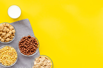 Various corn cereals in bowls and milk on yellow background top view copyspace
