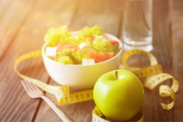 Salad in white bowl, fresh green apple and tape measure on dark wooden background. Diet and weight loss concept.