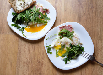 Fried eggs for breakfast with ham and arugula on wooden background, two dishes