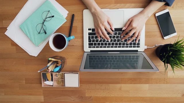 Top view of human female hands touching laptop keyboard typing in office working using internet. Smartphone, notebook and cup of coffee are visible on desktop.