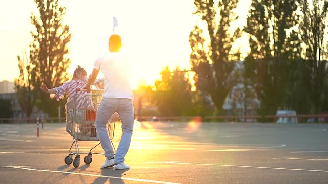 Young couple of blonde caucasian woman and man having fun at the car parking. Guy whirling girl in the supermarket cart at the parking. They are happy and laughing. Handheld shot.