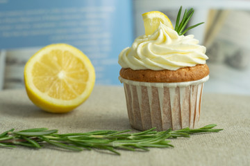 Homemade sweet lemon cupcakes with cheese cream, slice of lemon, branch of rosemary at blue cookbook background at linen tablecloth, selective focus, close up, copy space, from the top.
