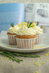 Homemade bakery lemon cupcakes with cheese cream, slice of lemon, branch of rosemary at blue cookbook background at linen tablecloth, selective focus, close up, copy space,