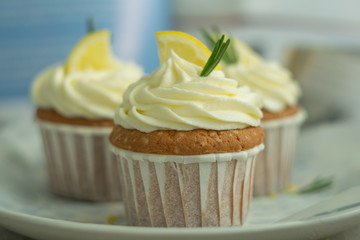 Fresh baked desserts lemon cupcakes at big plate with cheese cream, slice of lemon, branches of rosemary at cookbook blur background at perchament, selective focus, close up, copy space