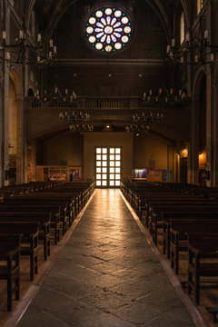 A dark indoor view of a late Gothic church. The aisle of the church, surrounded by banches in a row and at the background there is the main entrance door below a rose window.