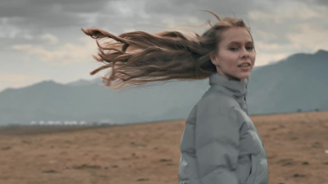 Close-up portrait: beautiful young girl with long hair in a jacket running in the open nature against the mountains, looking at the camera, spinning, hair develops wind. Cloudy light, slow motion, 4K