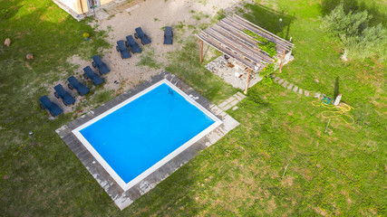 Aerial view of a blue pool with stairs to descend and climb into the water. Around marble tiles. There are empty loungers by the pool. The pool is part of a private villa.