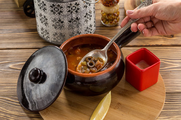 female hand holding spoon of traditional Russian soup solyanka with smoked meat in clay pot on wooden table
