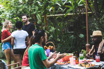 Group of people standing around barbecue grill, chatting, drinking and eating at summer outdoor party and holidays concept..