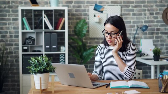 Female office worker talking on mobile phone and using laptop then taking notes writing in notebook working in loft style room. People and communication concept.