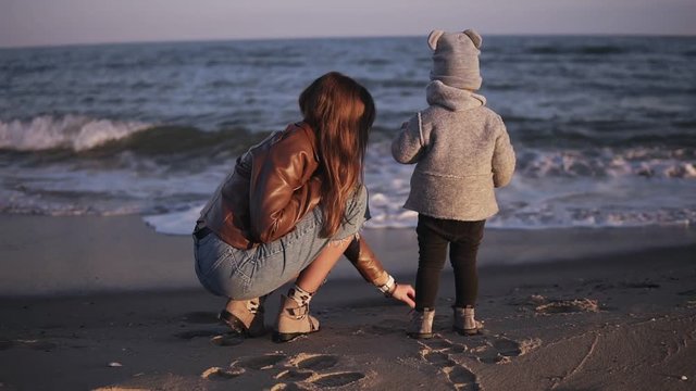 Backside footage of a mother and her child against the sea or ocean. Little, cute baby standing and looking on the water. Woman collecting shells and stones for child. Cold weather