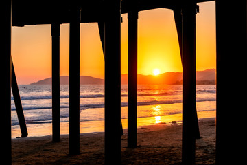 Orange sunset through Pylons of Pier at Beach over Ocean