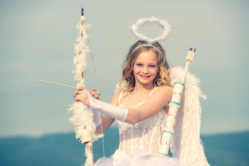 Innocent Girl with angel wings standing with bow and arrow against blue sky and white clouds. St...