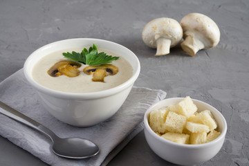 Champignon mushroom cream soup in a white bowl with parsley and crackers on a concrete gray background with copy space