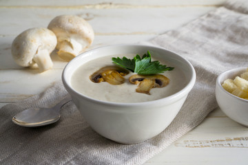 Champignon mushroom cream soup in a white bowl with parsley and crackers on a white wooden background with copy space
