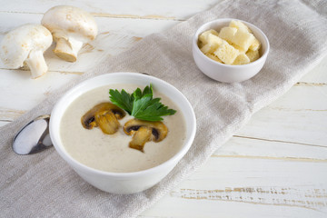 Champignon mushroom cream soup in a white bowl with parsley and crackers on a white wooden background with copy space