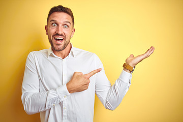 Fototapeta premium Young handsome man wearing elegant white shirt over yellow isolated background amazed and smiling to the camera while presenting with hand and pointing with finger.