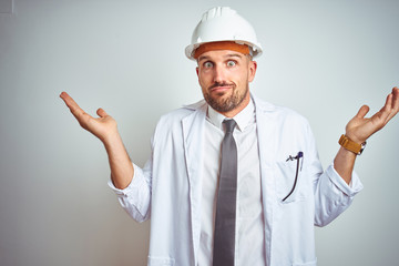 Young handsome engineer man wearing safety helmet over isolated background clueless and confused expression with arms and hands raised. Doubt concept.