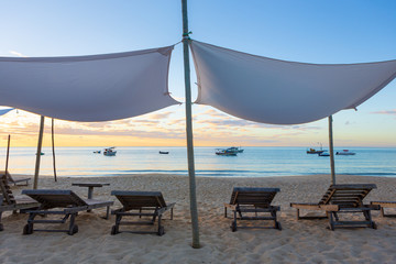 Beautiful beach with wooden chairs, sun loungers and tent with boats and sunset in the background.Concept of vacations, peace and relaxation. Ponta do Corumbau, Bahia, Brazil.