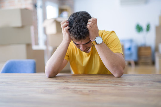 Young Man Sitting On The Table With Cardboard Boxes Behind Him Moving To New Home Suffering From Headache Desperate And Stressed Because Pain And Migraine. Hands On Head.
