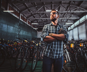 Obraz na płótnie Canvas Pensive man in protective glasses and checkered shirt is posing at his own warehouse full of bicycles.