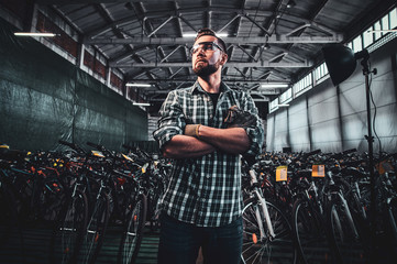 Fototapeta na wymiar Pensive man in protective glasses and checkered shirt is posing at his own warehouse full of bicycles.