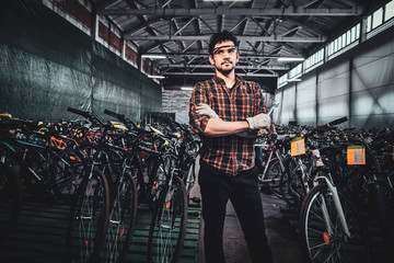 Attractive pensive mam in glasses is posing next to bicycles at his own warehouse.