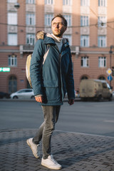 Vertical photo of handsome adult male in blue jacket with green backpack. Pretty man with beard and glasses walking on the street. Tourist exploring new city, travel. Cold season, autumn or winter. 