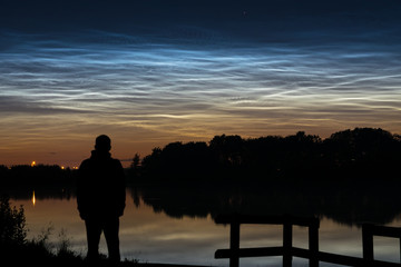 Fototapeta premium Silhouette of a person looking at noctilucent clouds (NLC, night clouds) near a lake in Holland in the evening.