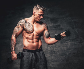 Athletic muscular shirtless male with tattoo on his chest doing biceps workouts with dumbbell.