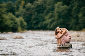 Young lonely girl sitting at stone in the river among mountains in summer.