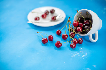 White cup with ripe berries of red sweet cherry and several berries in front of the cup. Composition on a blue background. red cherry berries in ceramic cup, autumn harvest.copy space