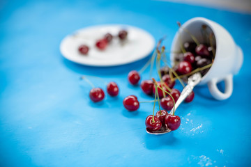 White cup with ripe berries of red sweet cherry and several berries in front of the cup. Composition on a blue background. red cherry berries in ceramic cup, autumn harvest.copy space
