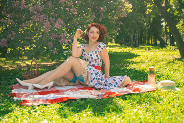 red-haired happy pin up girl in vintage summer dress and classic stockings with a seam in the back sits on a blanket  on the green grass in the city Park, next to a lilac Bush