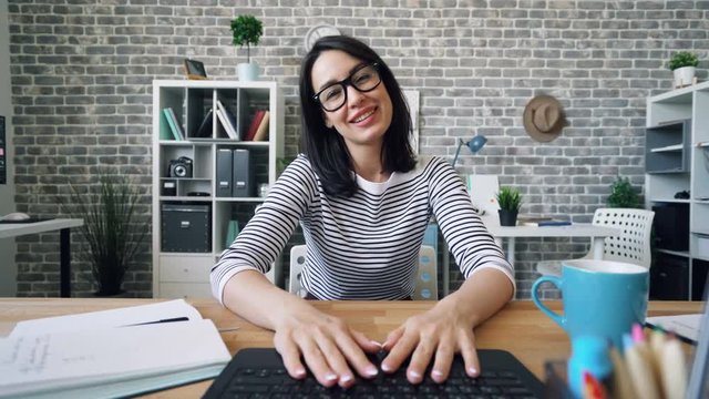 Portrait of attractive girl making video call with laptop in office skyping smiling gesturing waving hand sitting at desk and looking at camera. Communication and devices concept.