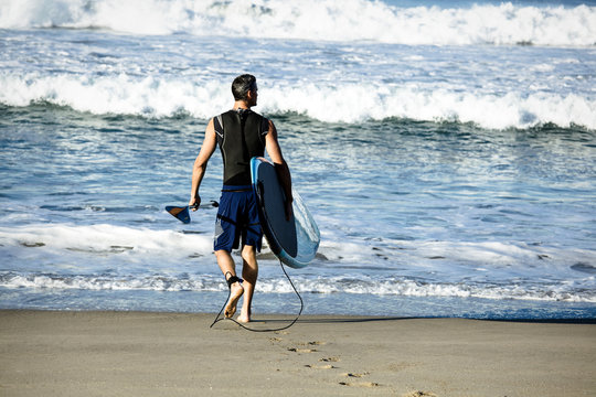 Surfer walks toward waves with paddle and surfboard - Powered by Adobe