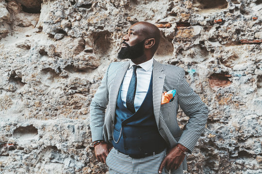 Handsome bald bearded black senior in an elegant costume in front of a grungy wall outdoors; a fashion adult African man with the beard and in a suit with vest and necktie standing near a stone wall