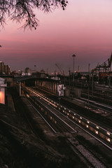 Vertical view of a modern railway depot during a lilac sunset in the dusk, with most of railroad tracks empty, with sequences of lights stretching into a hangar; port and dock area in the background