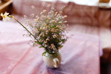 A vase of blue flowers with small buds on a table with a purple tablecloth against a rustic terrace with large Windows. Blurred background