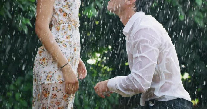 Slow motion of young handsome man is making a proposal of marriage to his fiancee under the rain on a background of green trees.