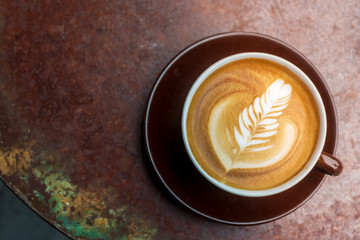 Top and close up view of a brown ceramic cup of cappuccino on top with beautiful delicate latte art with leaf pattern, and background of red rough rusty metal table. Vintage and industrial tone.