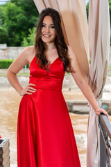 Fototapeta premium Young girl in red dress and teeth smile posing and holds a hand on hips in front of the iron fence and curtains in the garden on a summer day. Fashion and beauty concept. Close up, selective focus