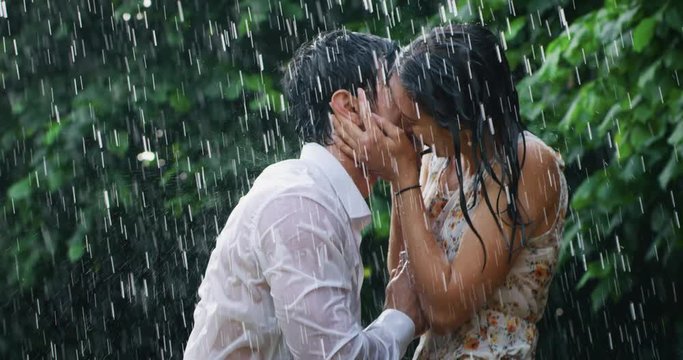 Slow motion of young carefree couple in love are hugging and kissing under the rain on a background of green trees.