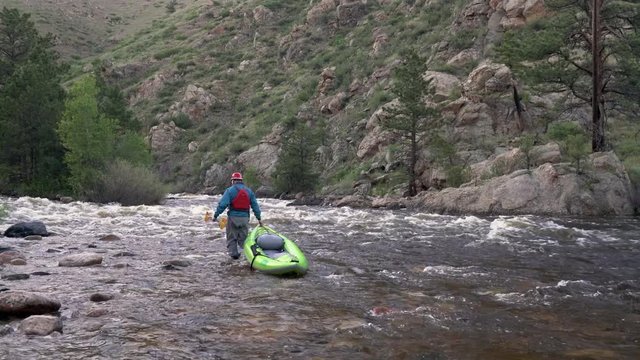 Male paddler is towing an inflatable whitewater kayak upstream of the mountain river, late spring scenery on the Poudre River in Colorado.