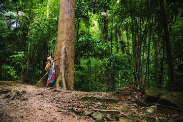 Naklejka na ściany i meble A girl with a backpack is embracing a big tree. enjoying travel nature in the forest Lush green trees. In the summer, Travel relax, Travel Thailand, tourist, countryside, outdoor, rain forest.
