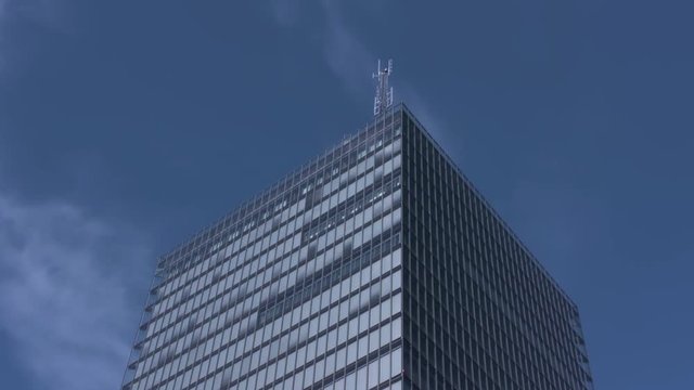 A light blinks on top of an antenna high atop a modern skyscraper. Bright blue sky and thin white clouds in the sky. Tall glass building reflects the daytime sun and sky giving a blue appearance.