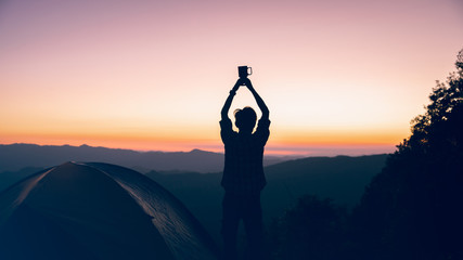 Hiker man holding coffee cup overhead near camping tent on mountains at sunset background. travel freedom time concept.