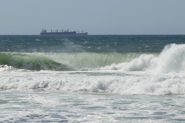 Big storm waves hitting the beach of the sea. Cargo ships at anchor on the background. Sunny day.