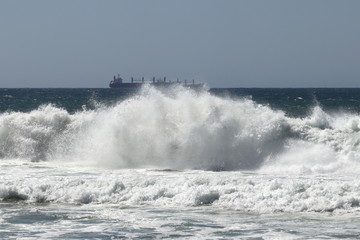 Big storm waves hitting the beach of the sea. Cargo ships at anchor on the background. Sunny day.