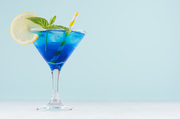 Summer cold blue lagoon drink with ice cubes, lemon slice, yellow straw, green mint in elegant wine glass on pastel mint background.